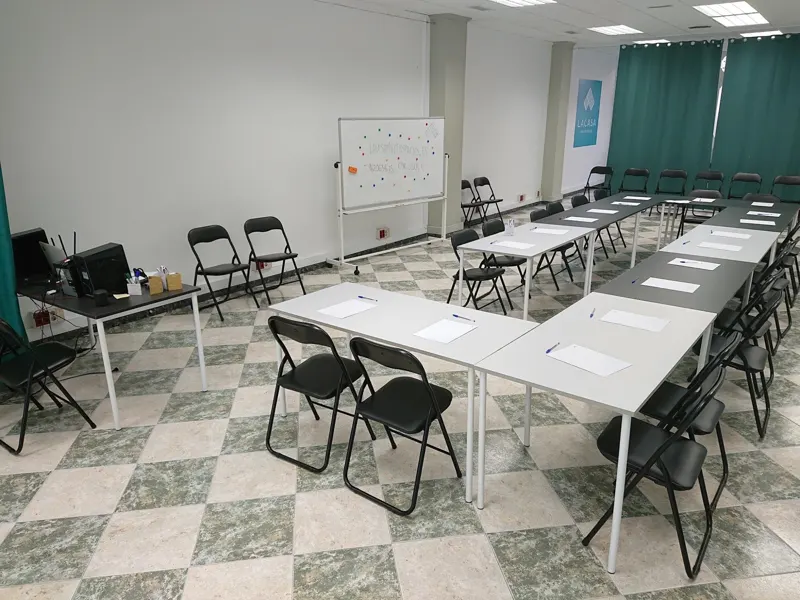 Mesas blancas en U con sillas plegables y una pizarra al fondo de la sala